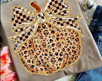 May include: A tan t-shirt with a pumpkin design. The pumpkin is decorated with a leopard print and skull accents. A large bow with a black and white checkered pattern sits atop the pumpkin. Gold glitter outlines the design.