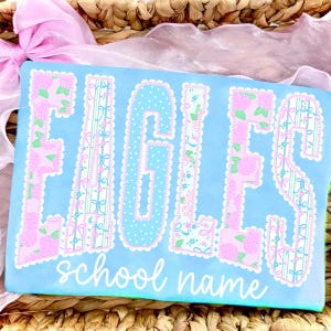 May include: A light blue rectangular item with the word "EAGLES" in large, decorative letters. The letters are adorned with floral and patterned designs in pink, green, and blue. Below the word is the text "school name" in a cursive font.