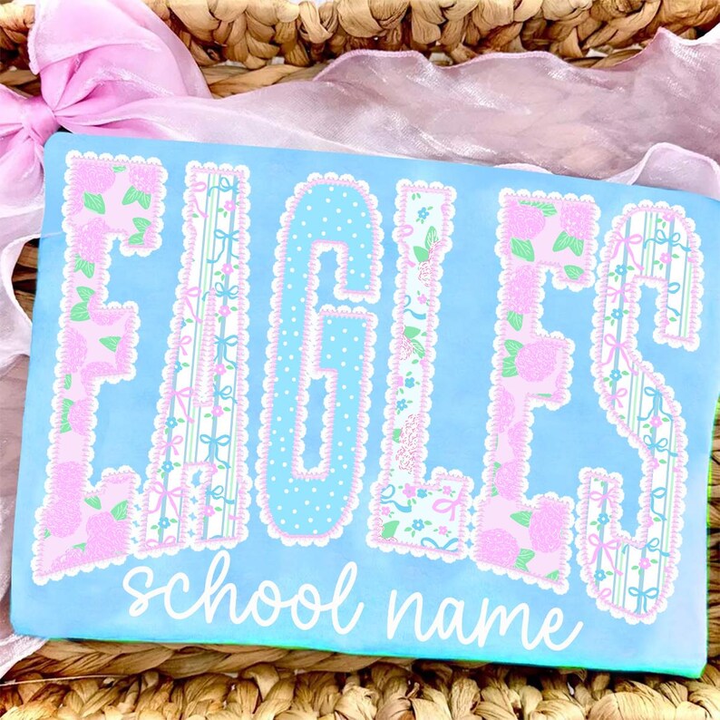 May include: A light blue rectangular item with the word "EAGLES" in large, decorative letters. The letters are adorned with floral and patterned designs in pink, green, and blue. Below the word is the text "school name" in a cursive font.