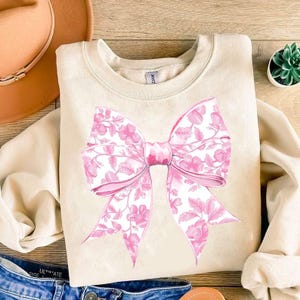 May include: A beige sweatshirt featuring a large pink floral bow design. The bow is centrally positioned on the sweatshirt.