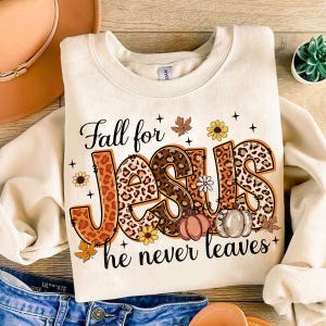 May include: A cream-coloured sweatshirt with the text "Fall for Jesus he never leaves" in black lettering. The word "Jesus" is decorated with a leopard print pattern and autumn-themed accents.