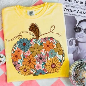 May include: Yellow Comfort Colors t-shirt with a floral pumpkin design. The pumpkin is filled with orange, blue, and pink flowers. The shirt has a crew neck and short sleeves. The text "COMFORT COLORS" is visible on the shirt.
