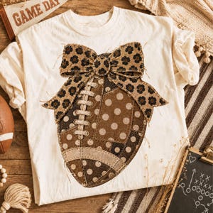 Könnte beinhalten: Ein cremefarbenes T-Shirt mit einer großen Grafik eines Footballs, der mit braunen und weißen Punkten sowie einer braunen Schleife mit Leopardenmuster verziert ist. Das Football-Design zeigt weiße Schnürsenkel und eine gesteppte Patchwork-Optik.