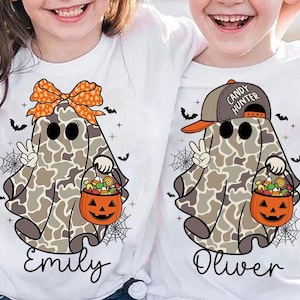 以下が含まれることがあります： ハロウィーンのゴーストデザインが施された2枚の白いTシャツ。1つのゴーストはリボンと「エミリー」という名前を身に着けています。もう1つのゴーストは「キャンディハンター」と「オリバー」という名前が書かれた帽子をかぶっています。各ゴーストは、キャンディーでいっぱいのジャック・オー・ランタンのバケツを持っています。