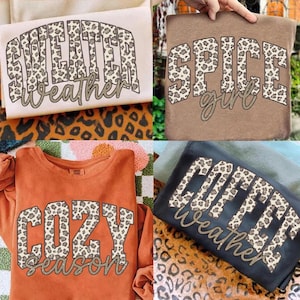 May include: Four t-shirts in cream, brown, orange, and black colours. Each shirt features a leopard print block letter design with cursive text underneath. The shirts display the phrases: Sweater Weather, Spice Girl, Cozy Season, and Coffee Weather.