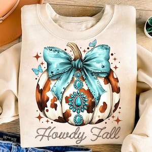 May include: A cream-coloured sweatshirt featuring a brown and white cow print pumpkin with a turquoise blue bow and rhinestones. The text "Howdy Fall" is written in brown rope-like letters below the pumpkin.