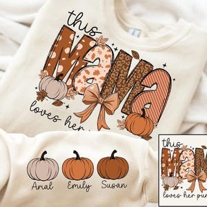 Può includere: Una felpa bianca con il testo "This Mama loves her pumpkins" in lettere marroni e arancioni. Il testo è circondato da un disegno di zucche e foglie. La felpa ha anche tre zucche più piccole con i nomi "Arial", "Emily" e "Susan" stampati sotto.