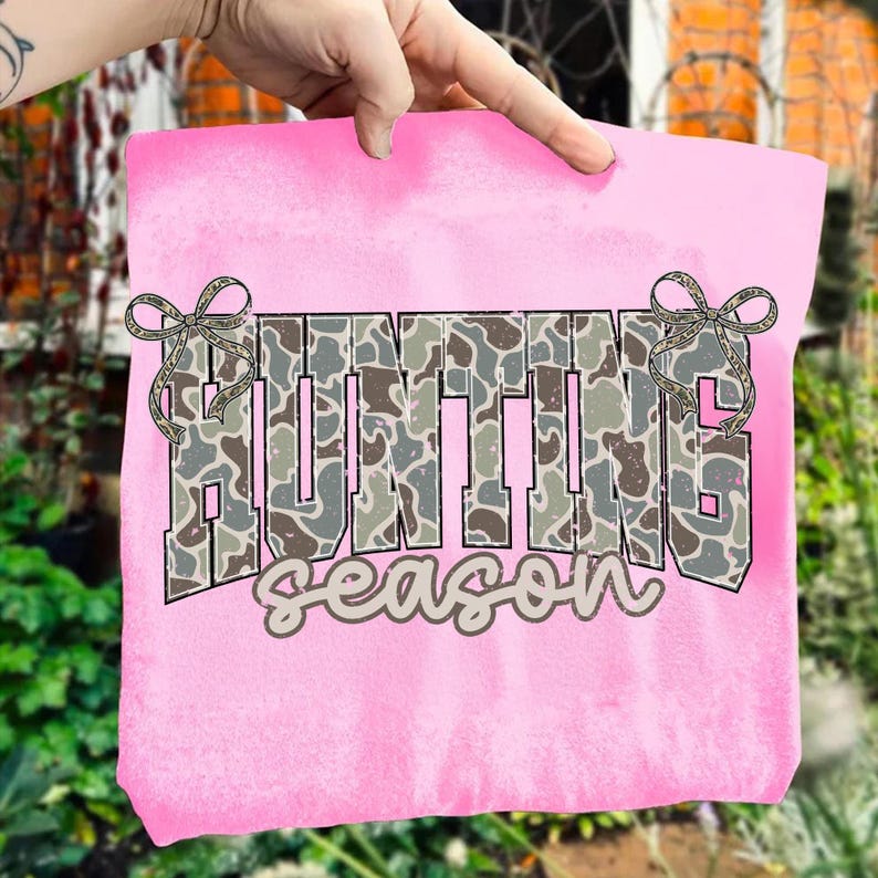 May include: A pink t-shirt with the words "HUNTING season" in a camouflage pattern. The text is outlined in black and has decorative bows on each side. The shirt is being held up by a hand.
