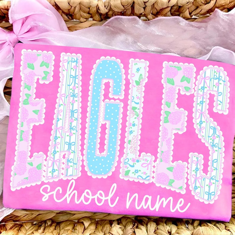 May include: A pink shirt with the word "EAGLES" in large, decorative letters. The letters are a mix of floral and polka dot patterns, with a light blue "G". The words "school name" are written in cursive below the word "EAGLES".