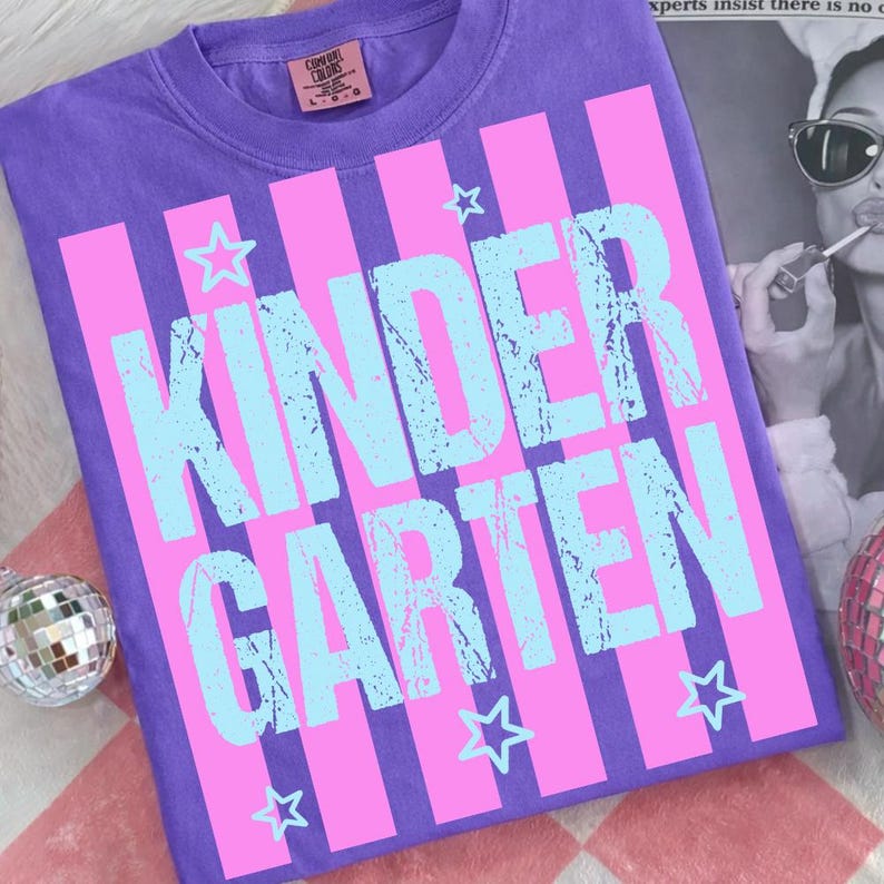 May include: A purple t-shirt with the word "KINDERGARTEN" in light blue, set against pink vertical stripes. Small white stars are scattered around the text. The shirt has a crew neck and a tag at the neckline.