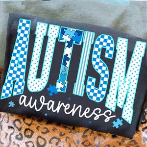 May include: A dark gray t-shirt with the word "AUTISM" in large, blue, and white letters. The letters have various patterns, including polka dots, puzzle pieces, and checkered designs. The word "awareness" is written in cursive below.