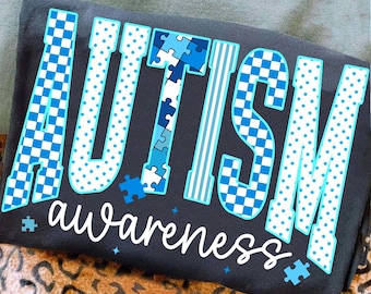 Op de afbeelding: Een donkergrijs t-shirt met het woord "AUTISM" in grote, blauwe en witte letters. De letters hebben verschillende patronen, waaronder stippen, puzzelstukjes en dambordontwerpen. Het woord "awareness" staat eronder in cursief.