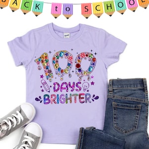 Op de afbeelding: Paars T-shirt met een glinsterend ontwerp met de tekst "100 dagen stralender" met een potlood en een gloeilamp.