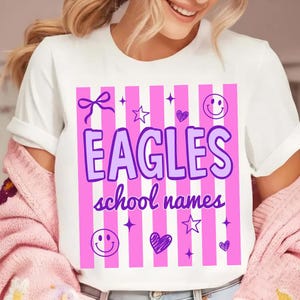 Op de afbeelding: Wit T-shirt met een roze en witte streepjesdesign. Het shirt heeft het woord "EAGLES" in paars, samen met de tekst "school names". Decoratieve elementen zijn sterren, harten, strikken en smiley gezichten.