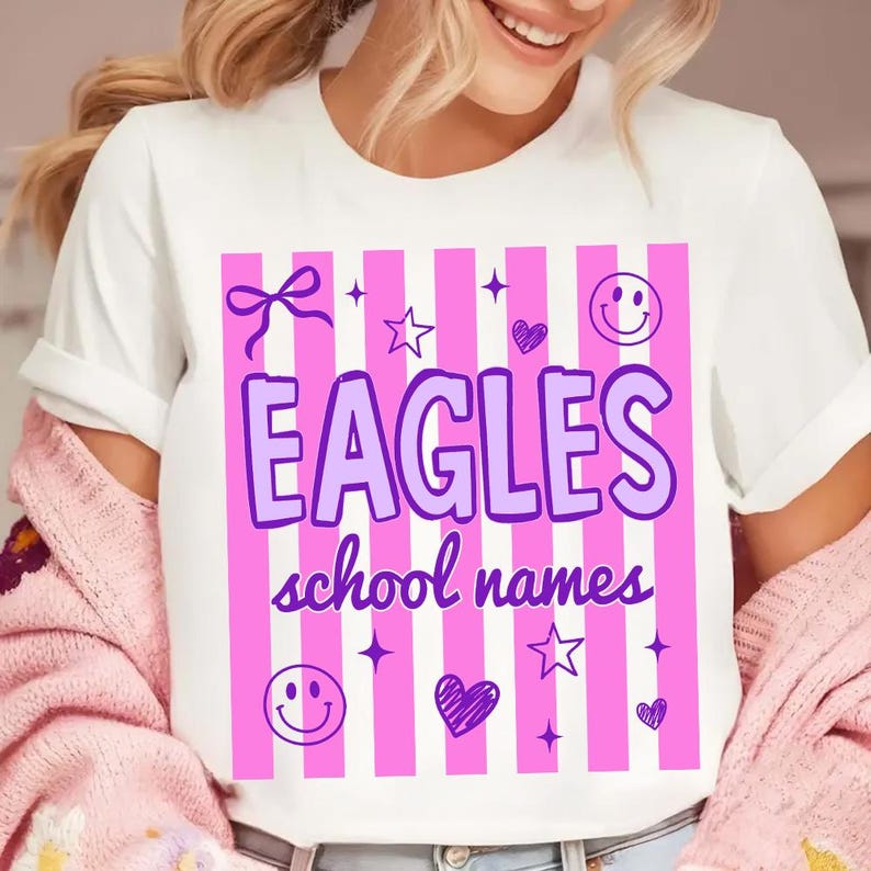 Op de afbeelding: Wit T-shirt met een roze en witte streepjesdesign. Het shirt heeft het woord "EAGLES" in paars, samen met de tekst "school names". Decoratieve elementen zijn sterren, harten, strikken en smiley gezichten.