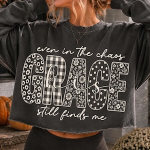 May include: A charcoal gray long-sleeve sweatshirt with a raw-edge hem. The front features the text "even in the chaos" above the word "GRACE" in large block letters with floral and plaid patterns, followed by "is still finds me" in script.