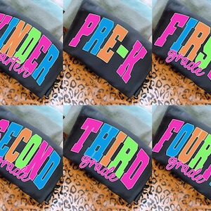 Puede incluir: Camisetas negras con texto colorido para diferentes grados escolares: Kindergarten, Pre-K, First Grade, Second Grade, Third Grade y Fourth Grade. El texto es rosa, naranja, verde y azul.