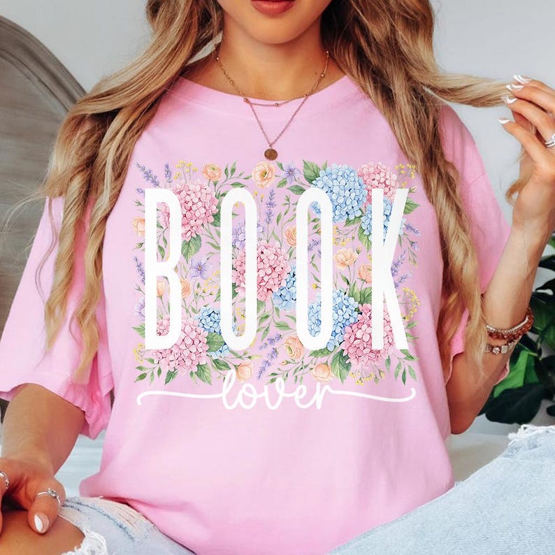 May include: A pale pink t-shirt featuring the text "BOOK lover" in white, framed by a floral arrangement of pink, blue, and purple blooms. The top has a relaxed fit.