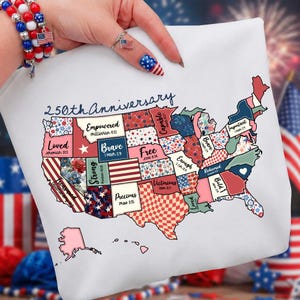 May include: White tote bag featuring a colorful map of the United States with each state decorated in patriotic patterns. The text "250th Anniversary" is above the map. The bag is held by a person wearing a bracelet and ring with American flag designs.