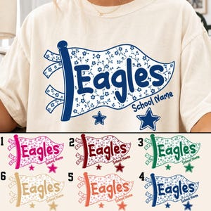May include: A cream-coloured t-shirt featuring a graphic of a pennant flag with a floral pattern and the word Eagles in blue. Below the flag, the text School Name appears alongside two blue stars. Six colour variations are shown below the main shirt.