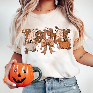 May include: A white t-shirt with the word "Teacher" in brown, orange, and white patterned letters. The letters are decorated with fall-themed elements like pumpkins, leaves, and a bow.