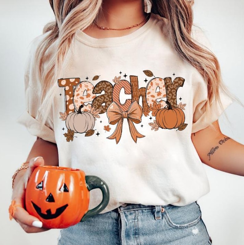 May include: A white t-shirt with the word "Teacher" in brown, orange, and white patterned letters. The letters are decorated with fall-themed elements like pumpkins, leaves, and a bow.