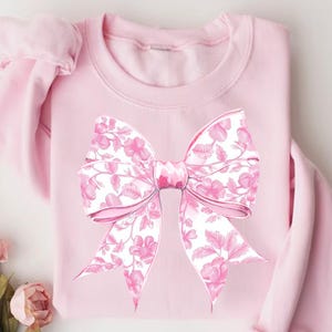 May include: A pink sweatshirt featuring a large pink bow printed on the front. The bow has a floral pattern in shades of pink and white.