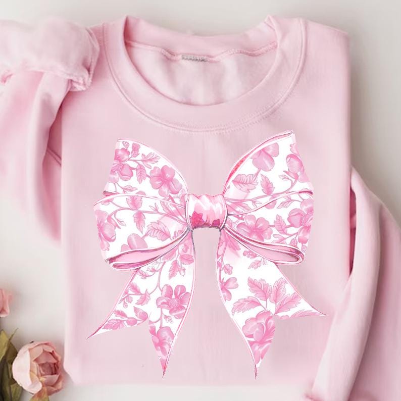 May include: A pink sweatshirt featuring a large pink bow printed on the front. The bow has a floral pattern in shades of pink and white.