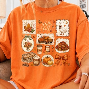 K&ouml;nnte beinhalten: Ein orangefarbenes T-Shirt mit einer Collage aus herbstlichen Illustrationen, darunter eine Gans, ein Lastwagen mit K&uuml;rbissen, ein Kaffeebecher, Zimtschnecken, eine Schleife und Bl&auml;tter. Der Text "Hello fall" ist mittig in Schreibschrift gedruckt.