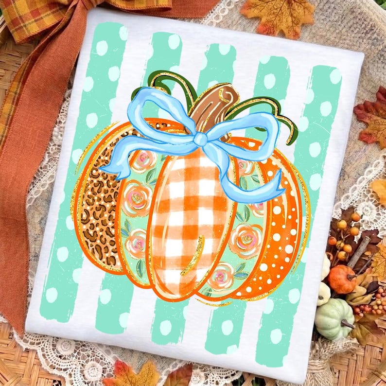 May include: A white t-shirt featuring a central pumpkin graphic with segments of leopard print, orange and white gingham, and floral patterns. A light blue bow sits atop the pumpkin, which is set against a background of mint green stripes.