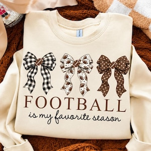 Könnte beinhalten: Ein cremefarbenes Sweatshirt mit Rundhalsausschnitt und drei dekorativen Schleifen auf der Brust. Die linke Schleife ist schwarz-weiß kariert, die mittlere hat ein Football-Muster und die rechte ist braun mit weißen Punkten. Der Text lautet FOOTBALL is my favorite season.