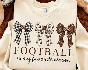 May include: A cream-colored crewneck sweatshirt featuring three decorative bows across the chest. The left bow is black and white checkered, the center bow has a football pattern, and the right bow is brown with white polka dots. The text reads FOOTBALL is my favorite season.