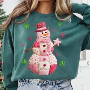 May include: Teal green sweatshirt with a patchwork snowman design. The snowman wears a top hat and scarf, holding a star wand. Pink and green snowflakes are scattered around the snowman, creating a festive winter theme.