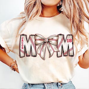 May include: A white t-shirt with a baseball-themed "MOM" design. The letters are made of baseballs with red stitching and a red bow in the center. The text "JACOB 18" is on the shirt.