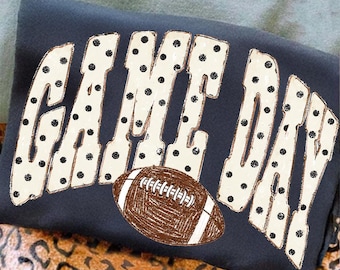 May include: A dark gray t-shirt featuring the text GAME DAY in large, cream-colored letters with black polka dots. A brown football with white laces and a textured, sketch-like appearance is positioned centrally below the text.