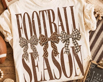 May include: A cream-colored t-shirt featuring the brown text "FOOTBALL SEASON" in a distressed font. Between the two words are three decorative bows: one with a leopard print, one with white polka dots on brown, and one with a brown gingham pattern.