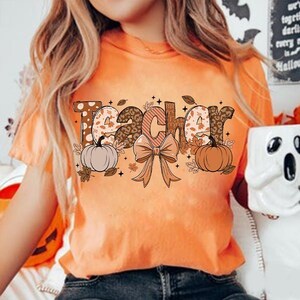 May include: Orange t-shirt with the word "Teacher" in brown, white, and black letters with a fall theme. The letters are decorated with polka dots, leopard print, and leaves. There are two pumpkins and a brown bow with leaves on either side of the word.