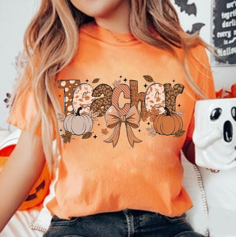 May include: Orange t-shirt with the word "Teacher" in brown, white, and black letters with a fall theme. The letters are decorated with polka dots, leopard print, and leaves. There are two pumpkins and a brown bow with leaves on either side of the word.