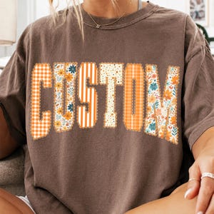 May include: A brown short-sleeve t-shirt featuring the word CUSTOM in large block letters across the chest. Each letter has a unique pattern including orange gingham, floral prints with blue and orange flowers, vertical orange stripes, and polka dots.