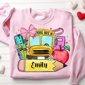 Pu&ograve; includere: Felpa rosa con un autobus scolastico, una matita, una mela, un righello e un fiocco. Il testo "Emily" &egrave; scritto sulla matita.