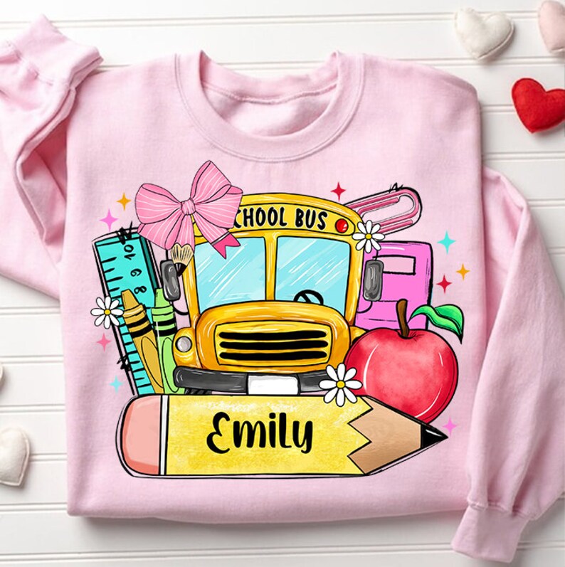 Pu&ograve; includere: Felpa rosa con un autobus scolastico, una matita, una mela, un righello e un fiocco. Il testo "Emily" &egrave; scritto sulla matita.