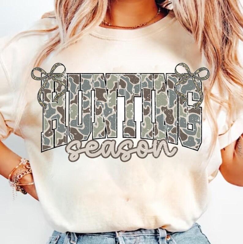 May include: A cream-colored t-shirt featuring the words "HUNTING season" in a camouflage pattern. The word "HUNTING" is in large, block letters, and "season" is in a script font. Decorative bows are on either side of the word "HUNTING".