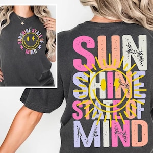 Puede incluir: Una camiseta gris oscura con un gráfico colorido en la espalda que dice "Sunshine State of Mind". El gráfico presenta un sol amarillo con una cara sonriente y texto rosa, naranja y morado.