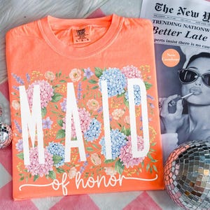 May include: Coral-colored t-shirt with the words "MAID of honor" in white script, surrounded by a floral design of pink, blue, and purple hydrangeas and other flowers. The shirt is on a pink and white checkered surface.