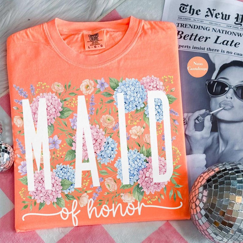 May include: Coral-colored t-shirt with the words "MAID of honor" in white script, surrounded by a floral design of pink, blue, and purple hydrangeas and other flowers. The shirt is on a pink and white checkered surface.
