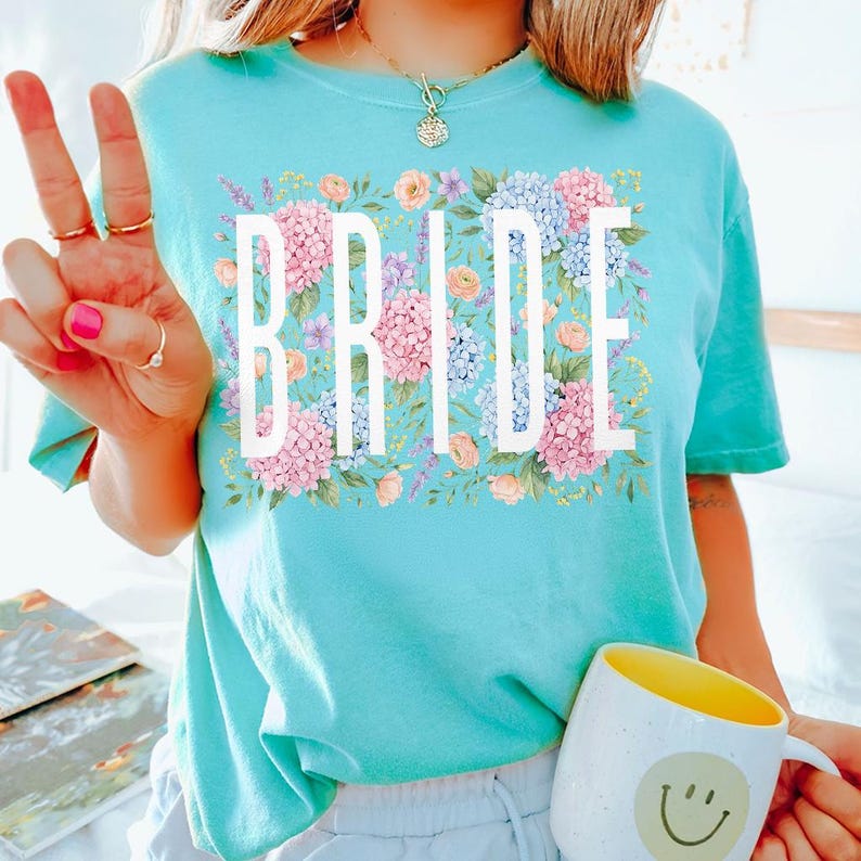 May include: A turquoise t-shirt with the word "BRIDE" in white block letters, surrounded by a floral design of pink, blue, and peach flowers. The shirt is a solid color and has a crew neck.