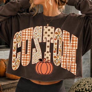 Könnte beinhalten: Ein dunkelbraunes bauchfreies Sweatshirt mit dem Wort CUSTOM in großen Blockbuchstaben. Die Buchstaben sind mit verschiedenen Mustern gefüllt, darunter orange-weißes Vichy-Karo, Blumenmuster und braune Punkte, mit einem orangefarbenen Kürbis darunter.