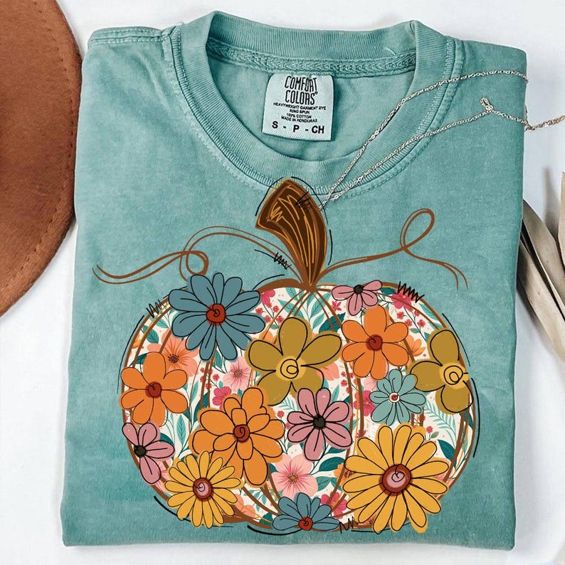 May include: A teal Comfort Colors t-shirt with a floral pumpkin design. The pumpkin is filled with orange, yellow, pink, and blue flowers. A silver necklace rests on the shirt. The shirt has a tag that says "Comfort Colors".