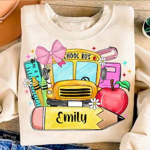 Pu&ograve; includere: Una felpa beige con un autobus scolastico, una matita, una mela, un righello e un fiocco. Il testo "SCHOOL BUS" &egrave; sull'autobus. Il testo "Emily" &egrave; sulla matita.
