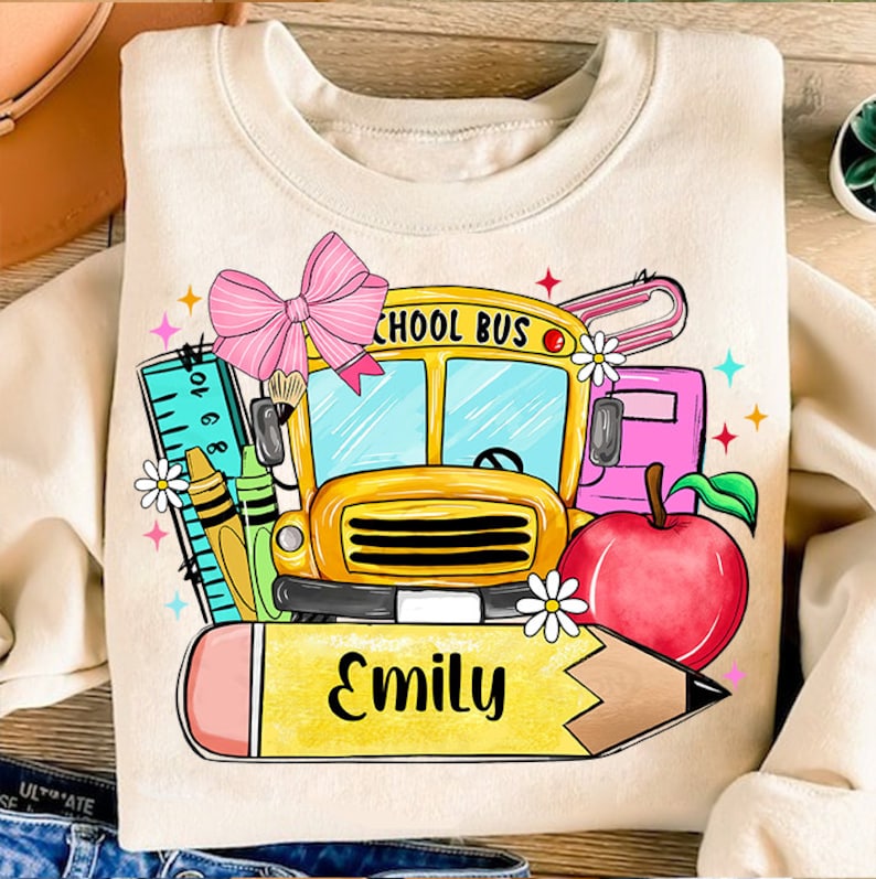 Pu&ograve; includere: Una felpa beige con un autobus scolastico, una matita, una mela, un righello e un fiocco. Il testo "SCHOOL BUS" &egrave; sull'autobus. Il testo "Emily" &egrave; sulla matita.